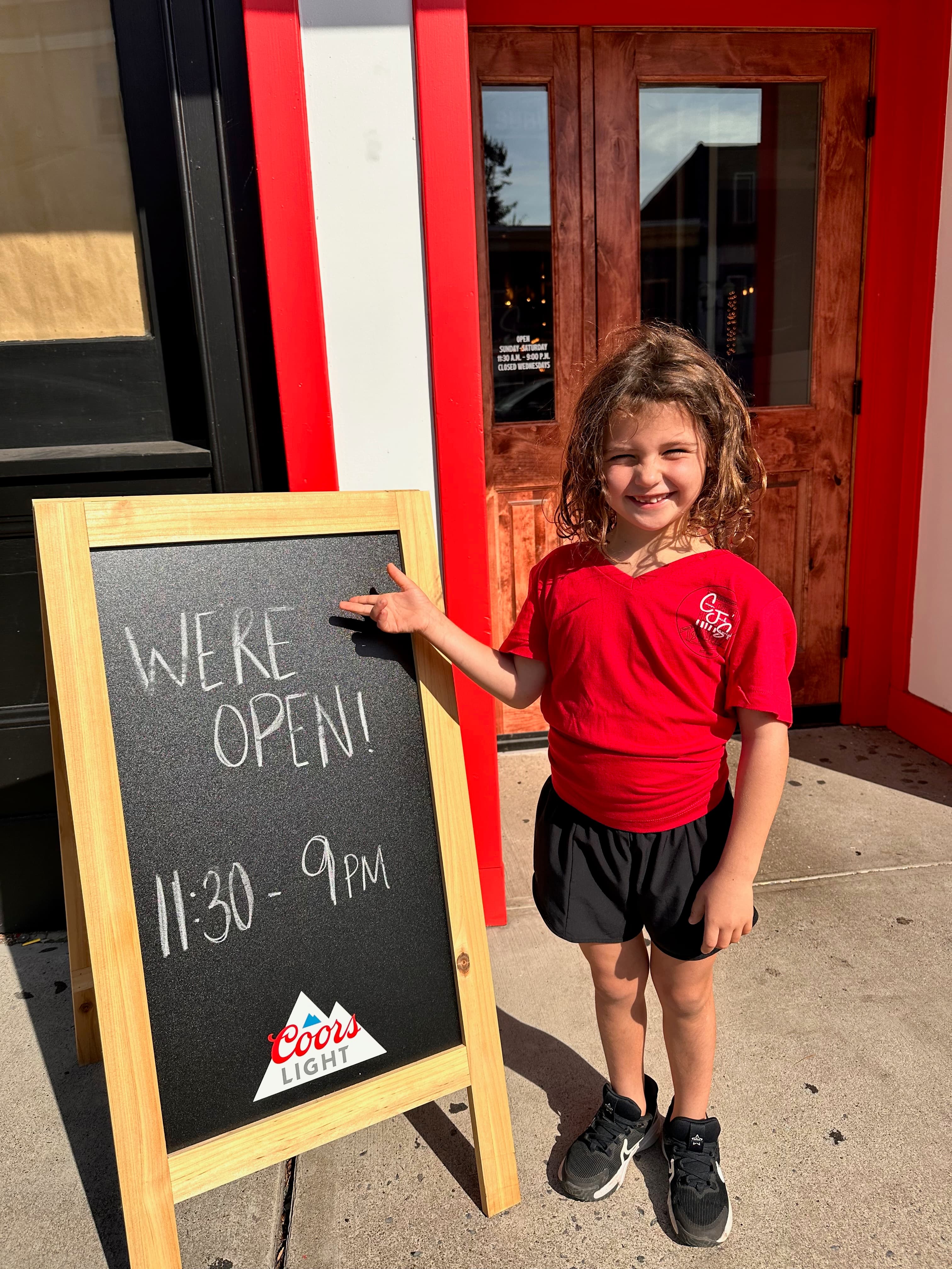 Young girl in red CJ'S shirt points to chalkboard sign reading 'WE'RE OPEN! 11:30 - 9PM'